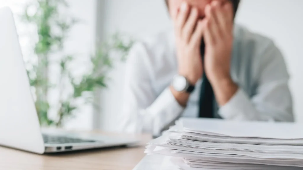 Preobremenjen moški z glavo v rokah sedi za pisalno mizo, obdano s kupom dokumentov – ponazoritev stresa in izgorelosti.
