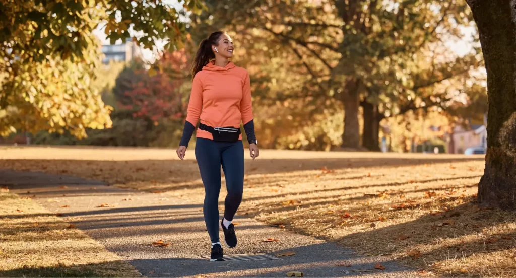 Ženska v oranžnem puloverju hodi po parku jeseni, nasmejana in sproščena – hoja v naravi kot podpora pri zmanjševanju tesnobe in paničnih napadov.