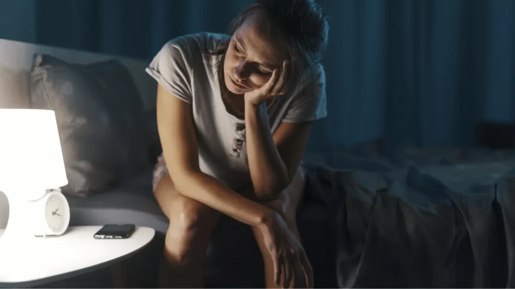 Ženska z nespečnostjo sedi na robu postelje in zaskrbljeno gleda v tla