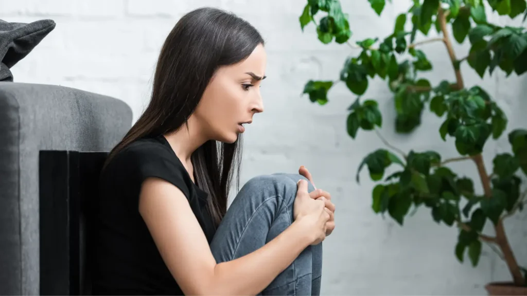 Ženska doživlja panični napad, sedi ob kavču, stisnjena vase in zaskrbljena, kar ponazarja simptome tesnobe.