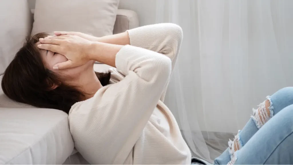 Ženska med paničnim napadom leži na tleh ob kavču, z rokami pokriva obraz in kaže znake tesnobe ter obupa.