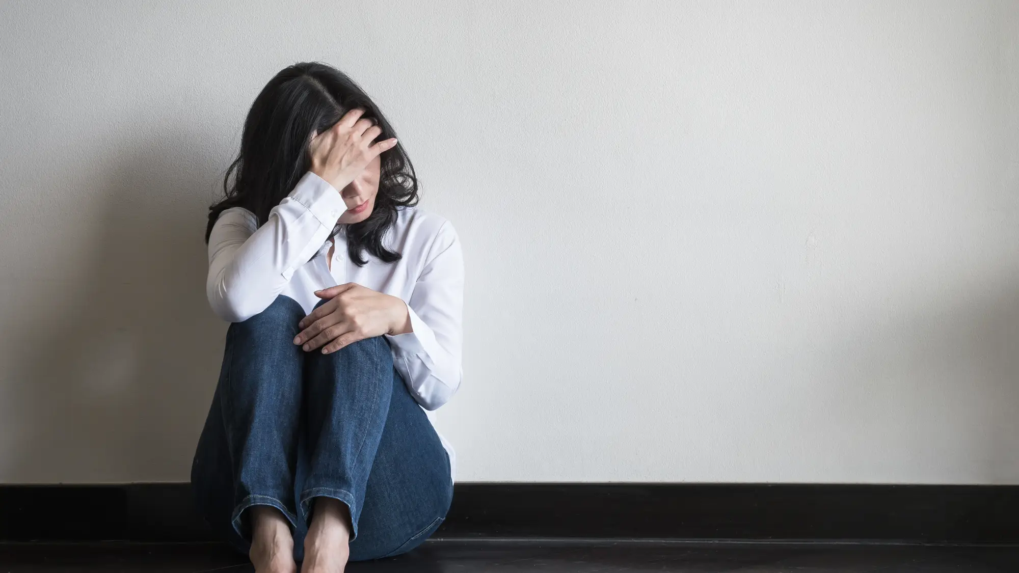 Ženska sedi na tleh ob steni z glavo naslonjeno na roko, izraz na obrazu kaže stisko in tesnobo - anksioznost.