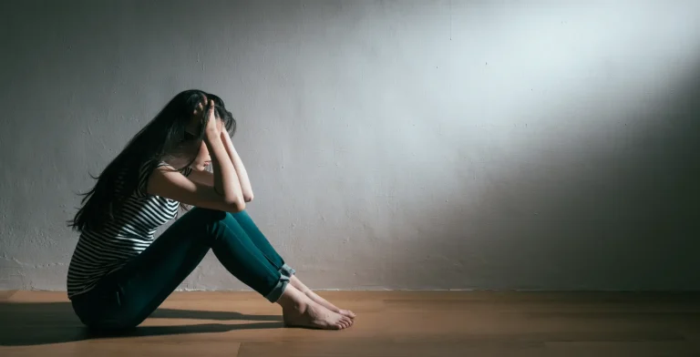 Mlada ženska sedi na tleh v temnem prostoru in si z rokami pokriva glavo – simbolični prikaz, kako se lahko kažejo znaki anksioznosti.