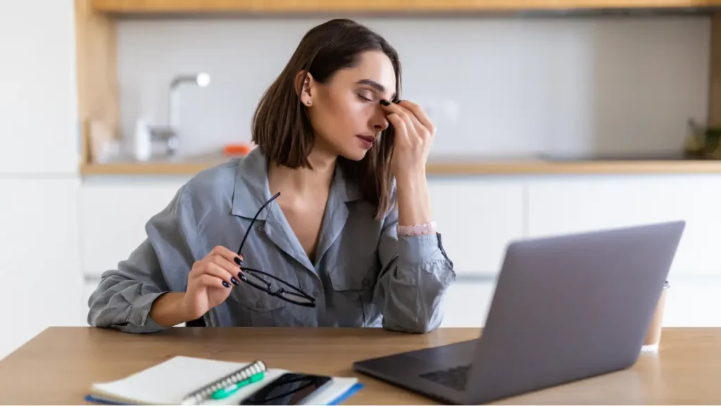 Ženska pred prenosnikom si z dlanjo pritiska ob oči zaradi utrujenosti in stresa pri delu